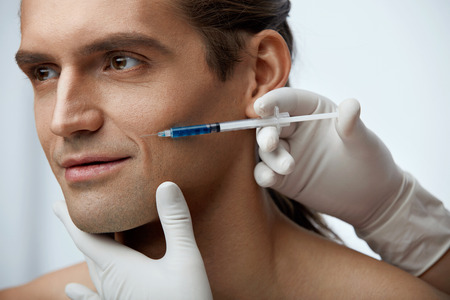 Beauty Injection. Portrait Of Handsome Sexy Man Getting Face Lifting Procedure On White Background. Close Up Doctor Hands Injecting Fillers Into Male Facial Skin. Cosmetology Concept. High Resolutionの写真素材