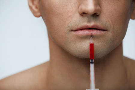 Facial Beauty. Closeup Of Handsome Man Face With Syringe With Red Liquid Near Lips. Male Patient Mouth Receiving Hyaluronic Collagen Injection Treatment. Lip Augmentation. High Resolutionの写真素材
