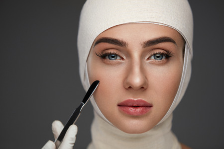 Plastic Surgery. Portrait Of Beautiful Young Woman With Smooth Soft Skin And Perfect Makeup. Closeup Of Attractive Female Patient With Head In Bandages And Scalpel In Hand. Skin Care. High Resolutionの写真素材