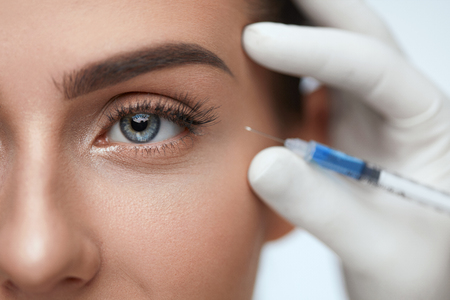 Face Lifting. Portrait Of Beautiful Woman's Face With Perfect Smooth Skin Receiving Hyaluronic Collagen Injection. Closeup Of Doctor Hands In Gloves Holding Syringe Near Woman Eye. High Resolutionの写真素材