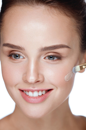 Woman With Beauty Face Applying Makeup. Portrait Of Beautiful Smiling Girl Putting Cream Foundation With Cosmetic Brush On Facial Skin. Closeup Sexy Young Female With Soft Smooth Skin. High Resolutionの写真素材