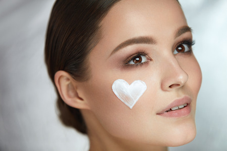 Woman Beauty Face Cosmetics. Closeup Sexy Smiling Female With Heart-Shaped Cosmetic Cream On Skin. Portrait Of Beautiful Happy Girl With Natural Makeup And Heart Sign On Soft Skin. High Resolutionの写真素材