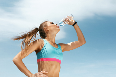 Woman Drinking Water After Running. Portrait Of Beautiful Athletic Girl In Bright Colorful Sportswear Resting After Fitness Workout, Drink Water From Bottle On Blue Sky Background. High Quality Imageの写真素材