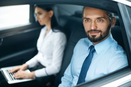 Portrait Of Business People In Car. Handsome Smiling Business Man In Elegant Shirt Riding To Work On Back Seat Of Luxury Car, Beautiful Woman On Background. Business Travel. High Quality Image.の写真素材