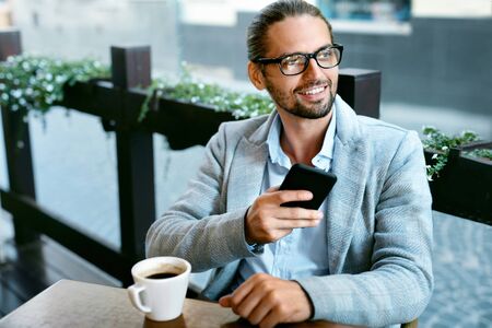 Man With Phone Drinking Coffee In Cafe. Portrait Of Handsome Smiling Young Businessman In Modern Men Clothes Using Mobile Phone, Enjoying Coffee Break Sitting At Table Outdoors. High Resolutionの写真素材