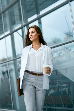 Beautiful Woman Going To Work With Coffee Walking Near Office Building. Portrait Of Successful Business Woman Holding Cup Of Hot Drink In Hand On Her Way To Work On City Street. High Resolution.の写真素材