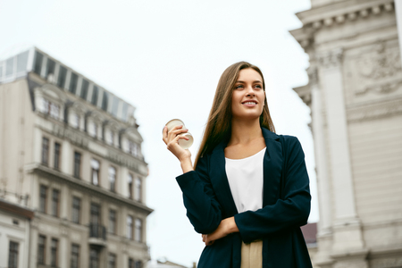 Beautiful Woman With Cup Of Coffee Walking On Street. Portrait Of Attractive Young Female In Stylish Office Clothes Holding Cup Of Hot Drink Standing Outdoors. High Resolution.の写真素材