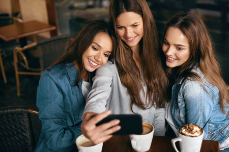 Friends Having Fun, Taking Photos In Cafe. Happy Gorgeous Young Women Taking Selfie Photos On Mobile Phone And Spending Leisure Time Together In Coffee Shop. Friendship. High Resolution.の写真素材