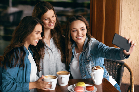 Beautiful Girls In Cafe Taking Selfie Photo On Phone. Happy Young Women Friends Having Fun, Taking Selfie Photos And Drinking Coffee In Coffee Shop. Friends Communication. High Quality Image.の写真素材