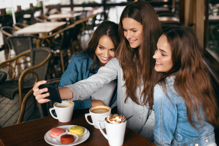 Beautiful Girls In Cafe Taking Selfie Photo On Phone. Happy Young Women Friends Having Fun, Taking Selfie Photos And Drinking Coffee In Coffee Shop. Friends Communication. High Quality Image.の写真素材