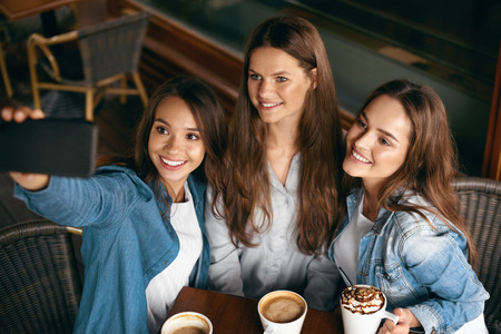 Friends Having Fun, Taking Photos In Cafe. Happy Gorgeous Young Women Taking Selfie Photos On Mobile Phone And Spending Leisure Time Together In Coffee Shop. Friendship. High Resolution.の写真素材