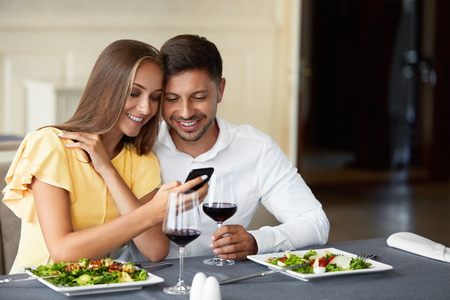 Couple In Love Looking On Phone Having Dinner In Restaurant. Lovely Young People Using Phone And Having Romantic Dinner In Restaurant. Relationships Concept. High Resolution.の写真素材