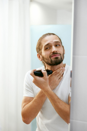 Man Shaving Face, Looking In Mirror. Portrait Of Young Attractive Shaving Beard With Trimmer In Bathroom. Men Face Hair Care. High Quality Image.の写真素材