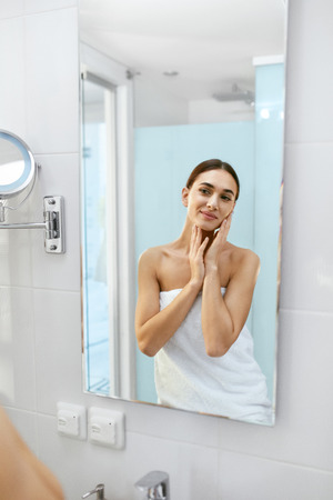 Happy Woman In Bathroom Looking In Mirror. Portrait Of Happy Female In White Towel On Body After Shower. Smiling Girl Touching Clean Soft Skin After Her Skincare Routine. High Resolution.の写真素材