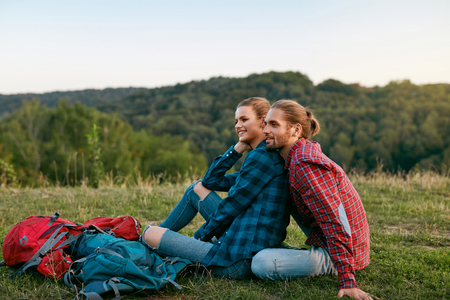 Young Tourist Couple Spending Time Together In Nature. Beautiful Smiling Couple In Love With Colorful Backpacks Relaxing While Sitting On Grass. Travel Concept. High Quality Image.の写真素材