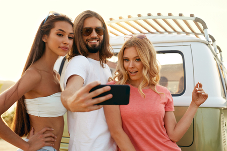 People Taking Photos With Phone During Summer Trip, Having Fun On Summer Travel Vacation. Smiling Young Friends Making Selfie On Mobile Phone Near Retro Bus Outdoors In Nature. High Resolution.の写真素材
