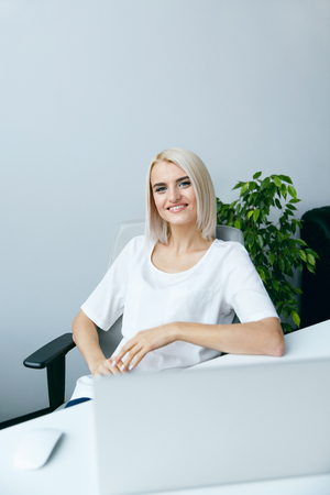 Young Woman Working On Notebook In Office. Portrait Of Beautiful Smiling Young Female Sitting At Desk, Working On Laptop At Workplace In Modern Light Office. Business People. High Resolution.の写真素材