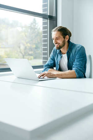 Man Working On Notebook In Office. Portrait Of Young Professional Smiling Man In Casual Clothes With Laptop Working At Workplace In Modern Light Office. Business People. High Qualityの写真素材