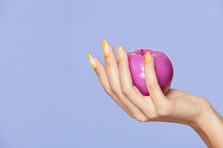 Nails Design. Female Hands With Colorful Nails Holding Apple On Violet Background. Close Up Of Woman Fingers With Fashion Orange Manicure Holding Purple Apple. High Quality Image.の写真素材