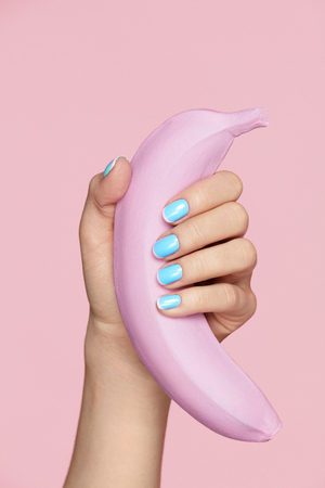 Nails Beauty. Hand With Blue Nails Holding Banana Fruit. Close Up Of Woman Fingers With Color Art Manicure And Pink Banana On Pink Background. Design For Nails. High Resolution.の写真素材