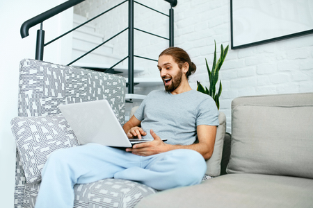 Young Man With Laptop At Home. Handsome Male Wearing Home Clothes Working At Computer, Sitting On Sofa. High Resolution.の写真素材
