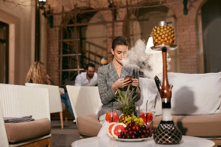 Woman With Phone Smoking Fruit Hookah In Shisha Bar. Girl Relaxing With Shisha. High Resolution