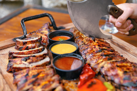 Meat Food. Barbecue Ribs And Sauces In Restaurant On Wooden Tray. Man's Hands With Ax Cutting Pork Ribs. High Resolutionの写真素材