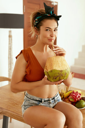 Healthy drink. Woman drinking coconut water in kitchen. Smiling young female in summer clothes...