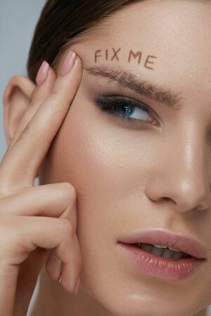 Beauty makeup. Woman face with messy eyebrow and fix me sign on skin closeup. Girl model with bushy fluffy messed up eyebrows and words written above. Eyebrow correction conceptの写真素材