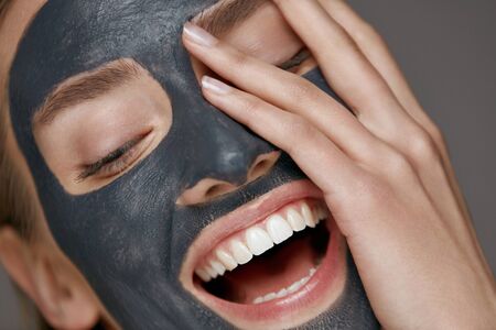 Beauty face skin care. Woman with cosmetic spa facial mask. Smiling girl model with grey clay mask and beautiful white smile closeup portraitの写真素材