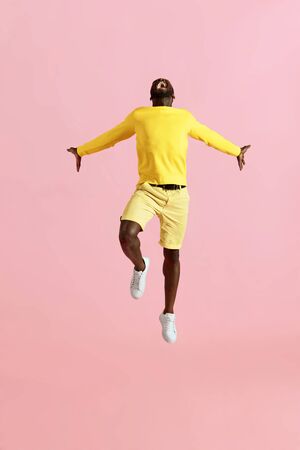 Jump. Black man jumping in air and screaming on pink background. Full length portrait of male model in yellow fashion clothes shouting while jumping at studioの写真素材