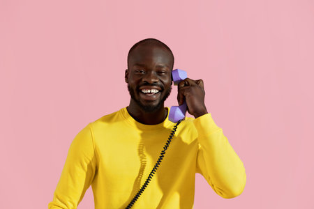 Smiling black man with purple phone on pink background colorful portrait. Happy young african american male model in yellow fashion clothes with purple retro corded phone in studioの写真素材