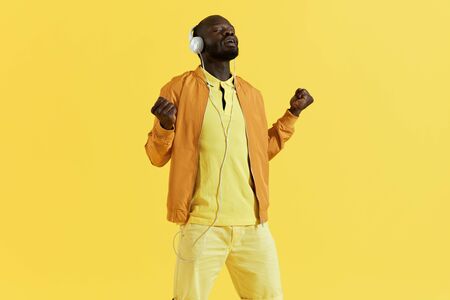 Man in headphones listening music, singing to song on yellow background. Portrait of black male model in white earphones and orange fashion clothes enjoying to singの写真素材