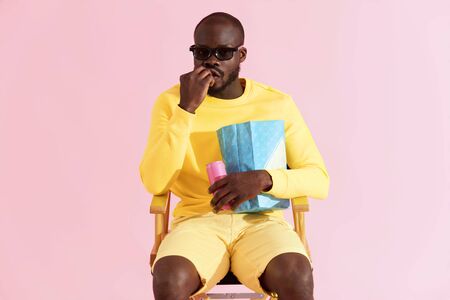 Man watching movie in cinema glasses eating popcorn portrait. Black male model in 3d glasses and yellow clothes sitting on chair watching show on pink background in studioの写真素材