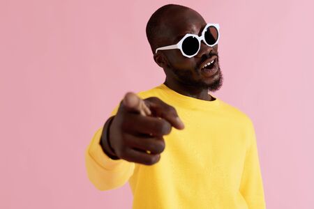 Fashion. Happy black man in sunglasses and yellow sweatshirt on pink background. Colorful portrait of smiling african american male model in glasses and hand pointing in cameraの写真素材