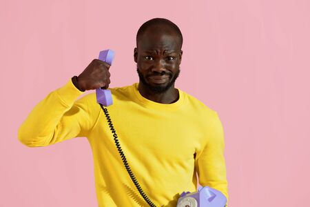 Black man having unpleasant phone call on pink background. Portrait of male with weird "yikes" face expression having uncomfortable talk on purple landline phone in studioの写真素材