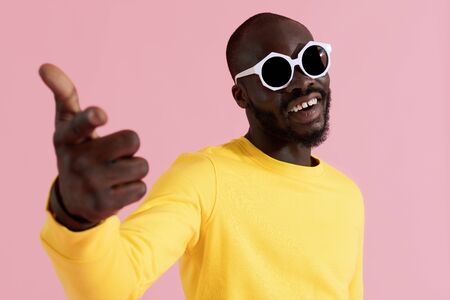 Fashion. Happy black man in sunglasses and yellow sweatshirt on pink background. Colorful portrait of smiling african american male model in glasses and hand pointing in cameraの写真素材