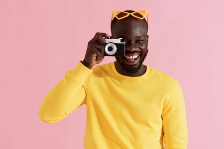 Happy man photographer with photo camera on colorful pink background. Portrait of smiling black male model in yellow fashion clothes taking photos in studioの写真素材
