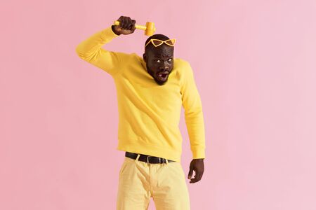 Funny black man beating his head with hammer on yellow background. Colorful portrait of african american male model in yellow fashion clothes behaving silly with mallet in studioの写真素材