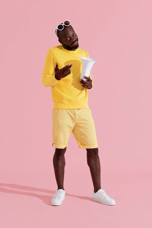 Black man in yellow fashion clothes with megaphone on pink background. Portrait of young male model with horn speaker in studioの写真素材