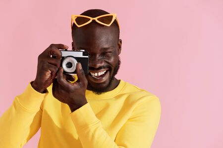 Happy man photographer with photo camera on colorful pink background. Portrait of smiling black male model in yellow fashion clothes taking photos in studioの写真素材