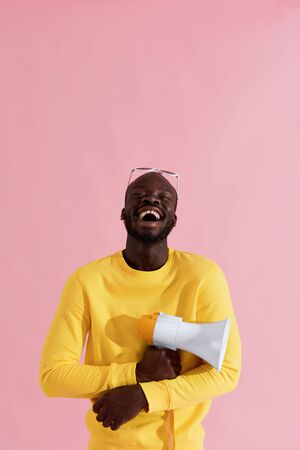 Happy black man in yellow fashion clothes with megaphone on pink background. Portrait of cheerful young male model with horn speaker laughing in studioの写真素材