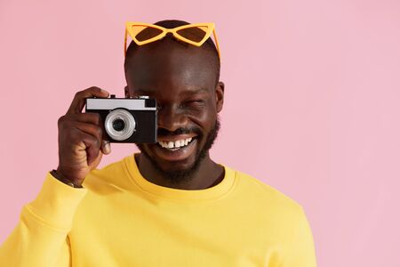 Happy man photographer with photo camera on colorful pink background. Portrait of smiling black male model in yellow fashion clothes taking photos in studioの写真素材