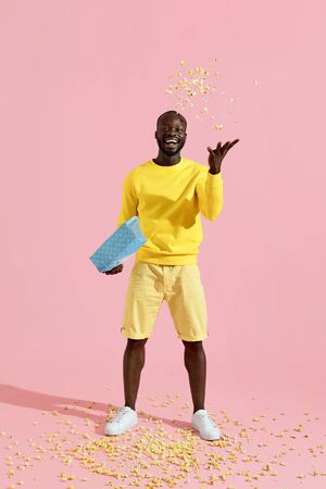 Happy black man having fun throwing pop corn in air. Full length portrait of cheerful male model in yellow clothes tossing popcorn on pink backgroundの写真素材