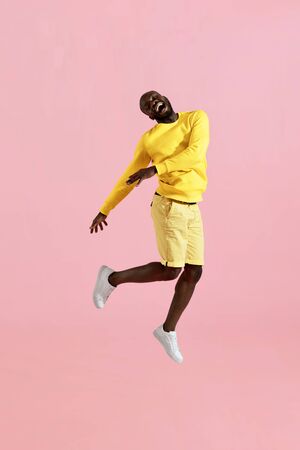 Jump. Happy black man jumping in air and laughing on pink background. Full length portrait of cheerful smiling male model in yellow fashion clothes having fun, studio shotの写真素材