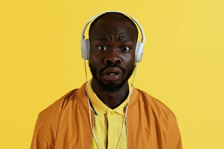 Surprised man in headphones listening music on yellow background. Portrait of amazed black male model with shocked face expression in white headsetの写真素材