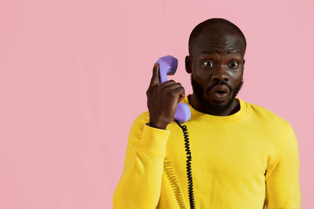 Surprised black man talking on retro phone on pink background. Colorful portrait of amazed african american male model with shocked face expression calling on purple landline phoneの写真素材