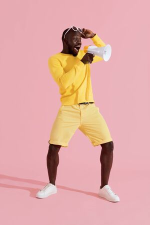 Attention! Black man shouting in megaphone on pink background. Full lenght portrait of happy young male model announcing in horn speaker in studioの写真素材