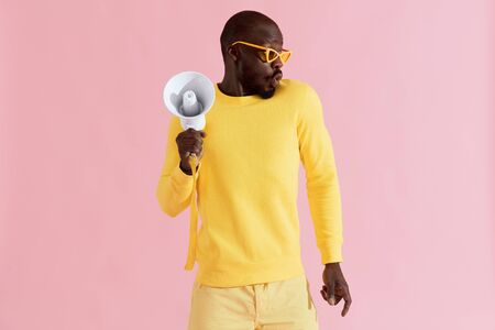 Black man in yellow fashion clothes with megaphone on pink background. Portrait of young male model with horn speaker in studioの写真素材