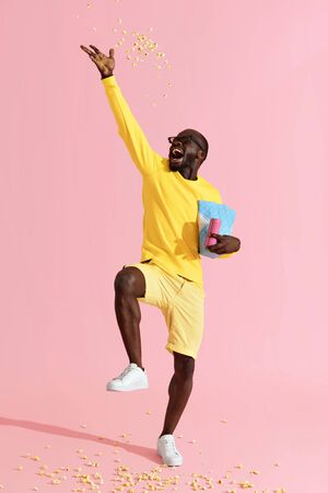 Happy black man in cinema glasses having fun throwing pop corn in air. Full length portrait of cheerful male model in movie glasses and yellow clothes tossing popcorn on pink backgroundの写真素材